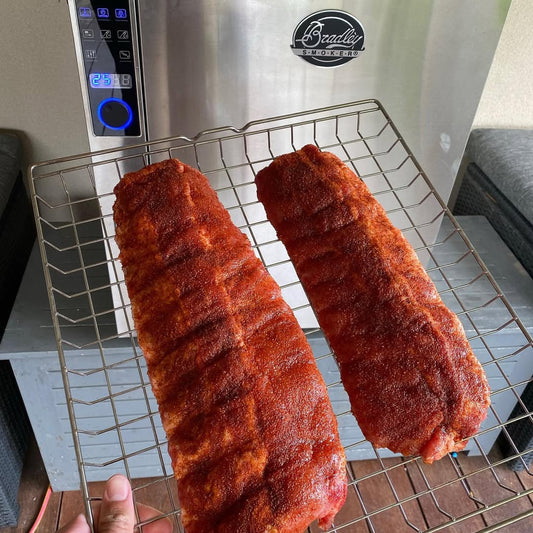 Two seasoned pork shoulders on a wire rack in front of a Bradley Smoker.