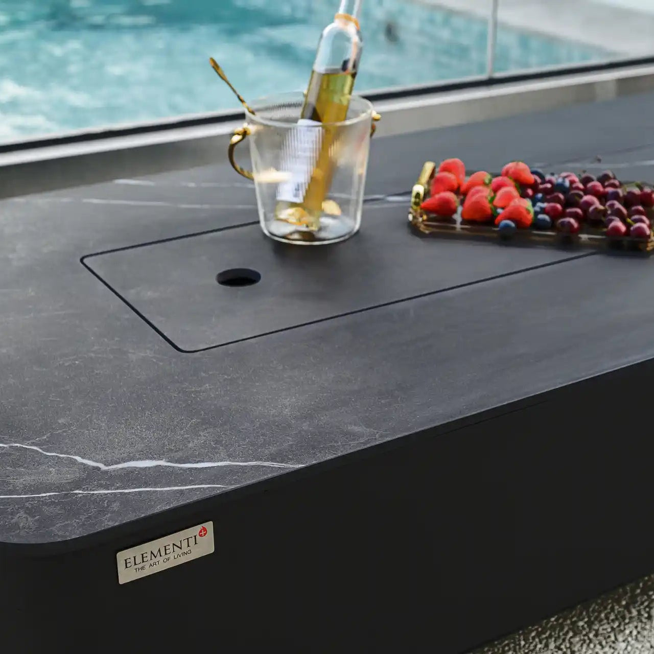 Outdoor table with a glass of champagne, fruit platter, and bottle of wine by a pool.