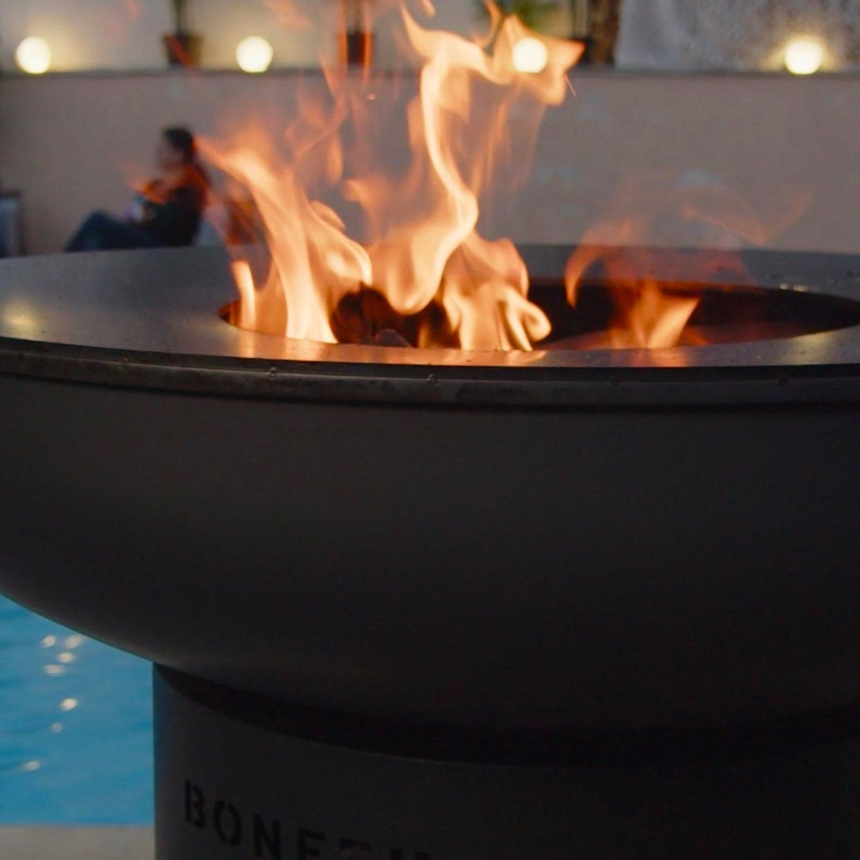 Fire pit with flames lit, featuring the brand 'BONFEU', near a poolside setting.