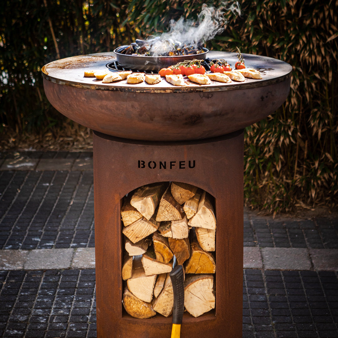 Rust-coloredBonfeu fire pit with wood storage and grill on a tiled patio.