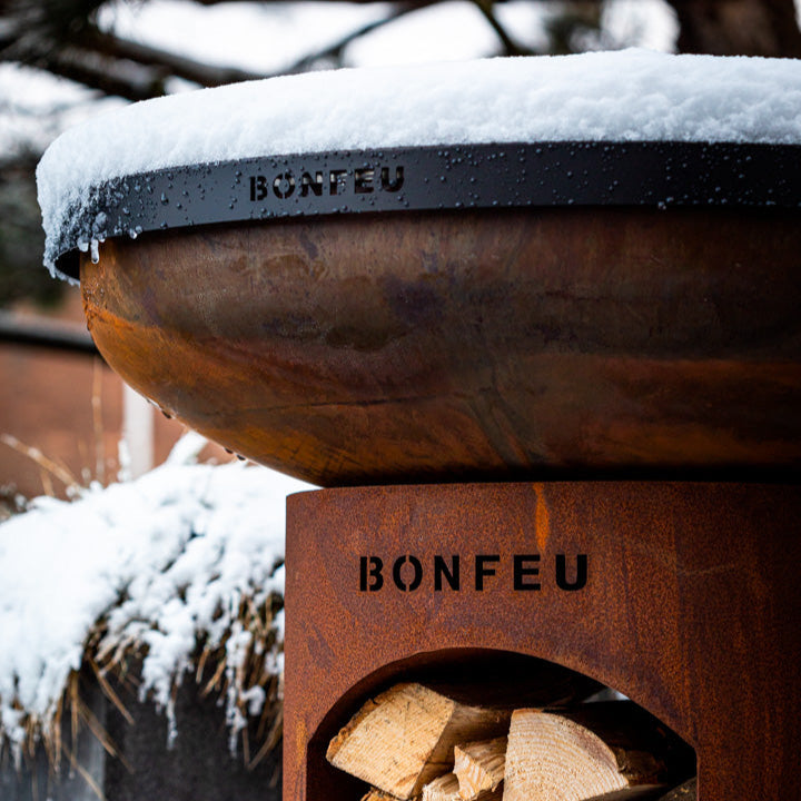 Wooden firewood holder with stacked logs and 'BONFEU' branding, covered in snow.