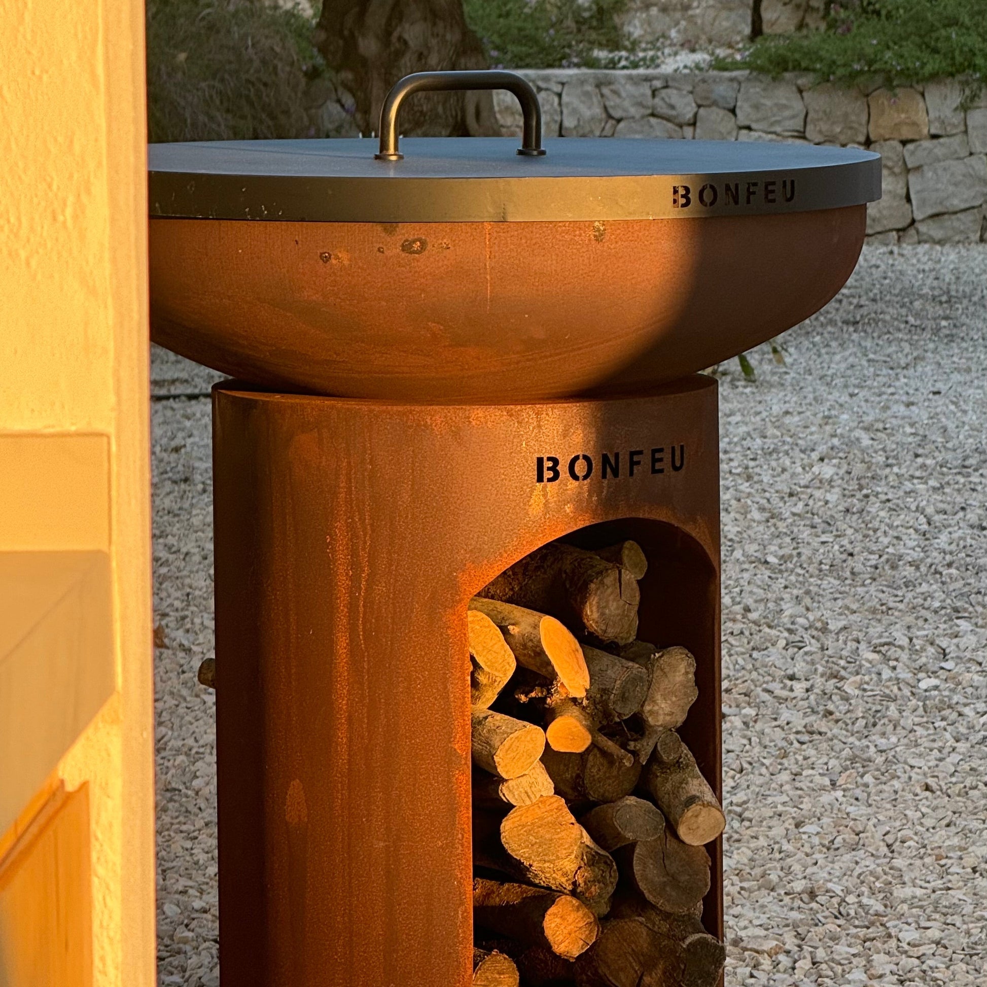 Rust-colored outdoor fire pit with wood storage on a gravel surface.