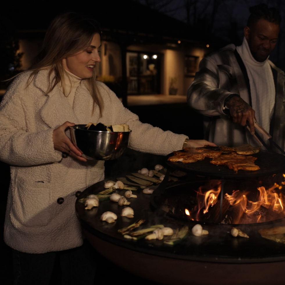 Two people cooking outdoors by a fire pit at night.