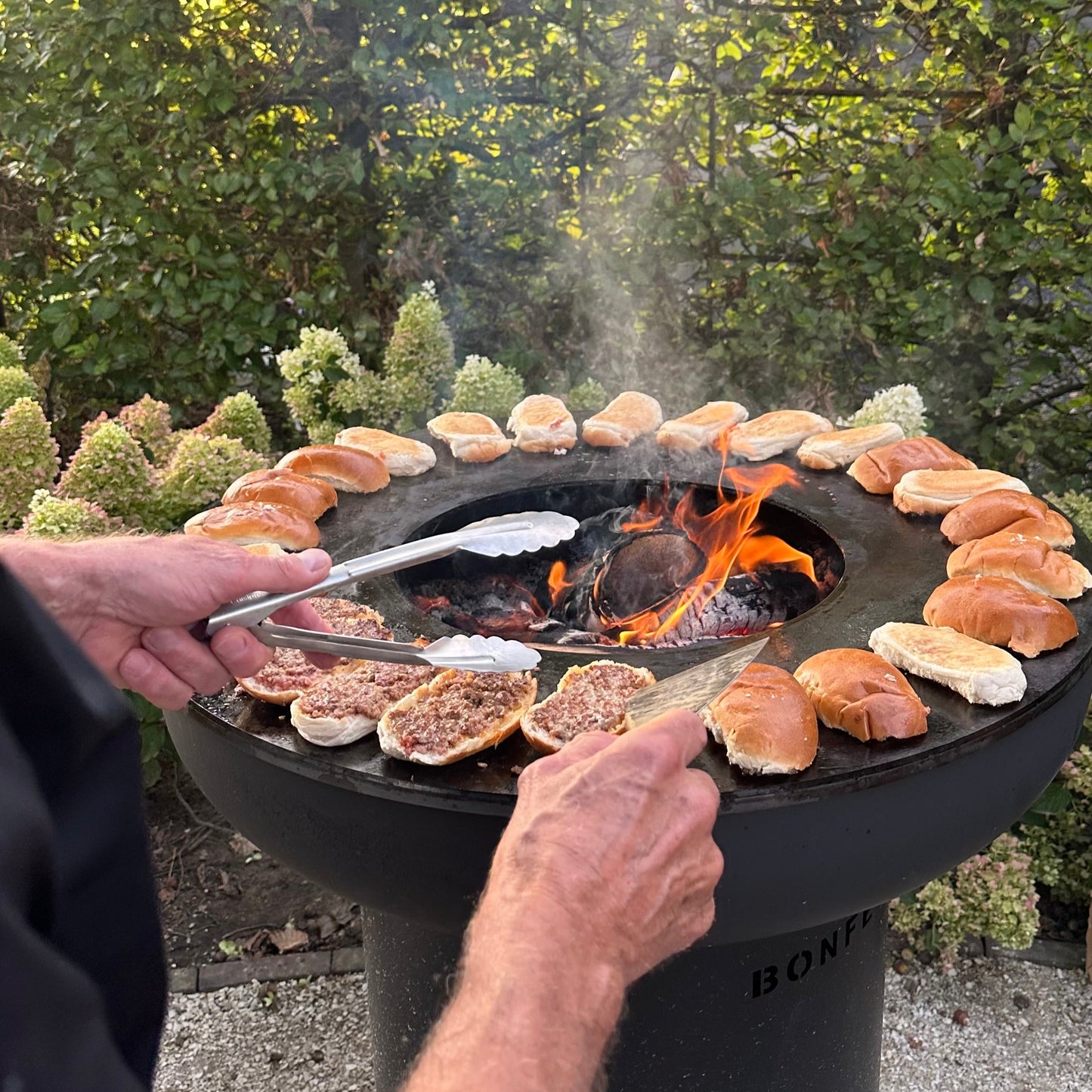 Person grilling bread buns on a portable grill outdoors with trees in the background