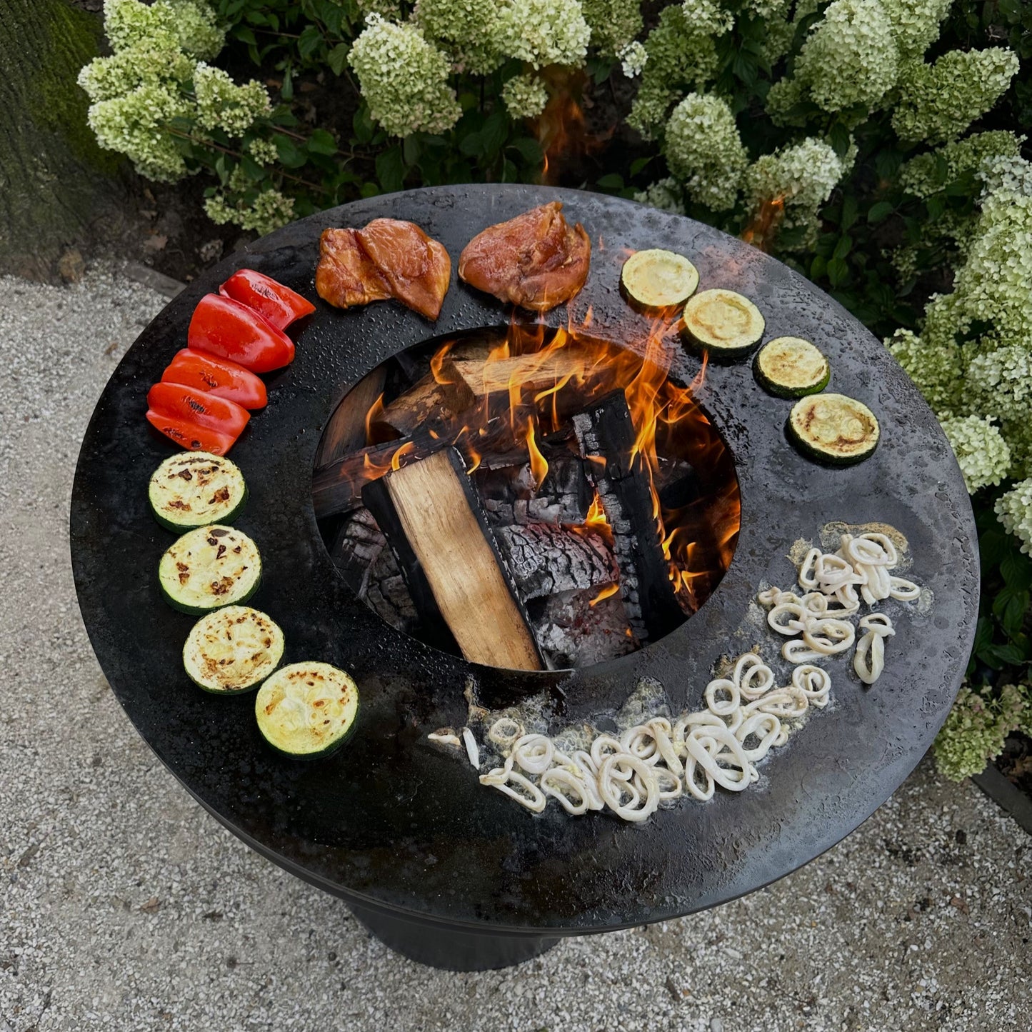 Fire pit with food items around it, surrounded by greenery