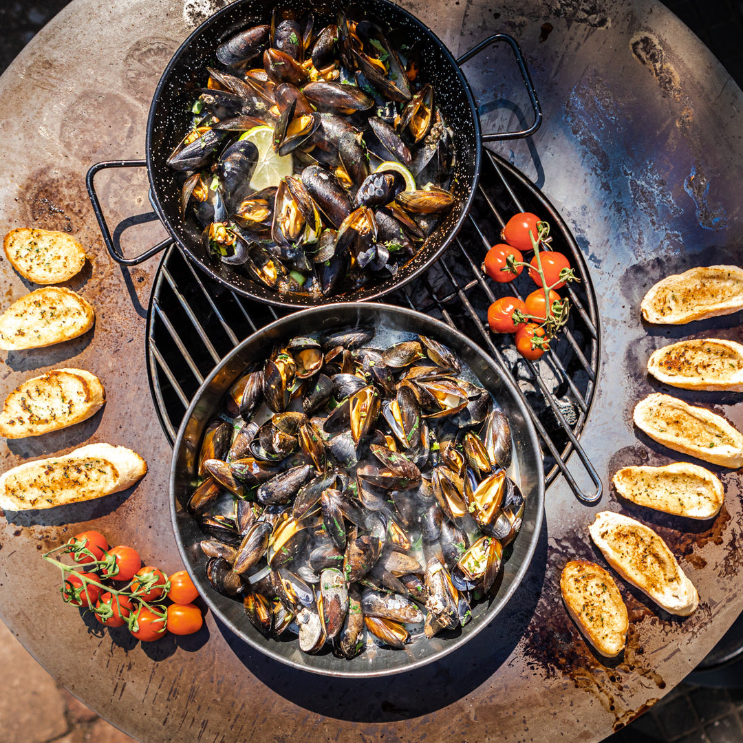 Two pans of mussels with bread and tomatoes on a stone surface