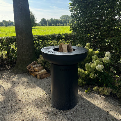 Black outdoor fire pit with wood logs in a garden setting with trees and fields