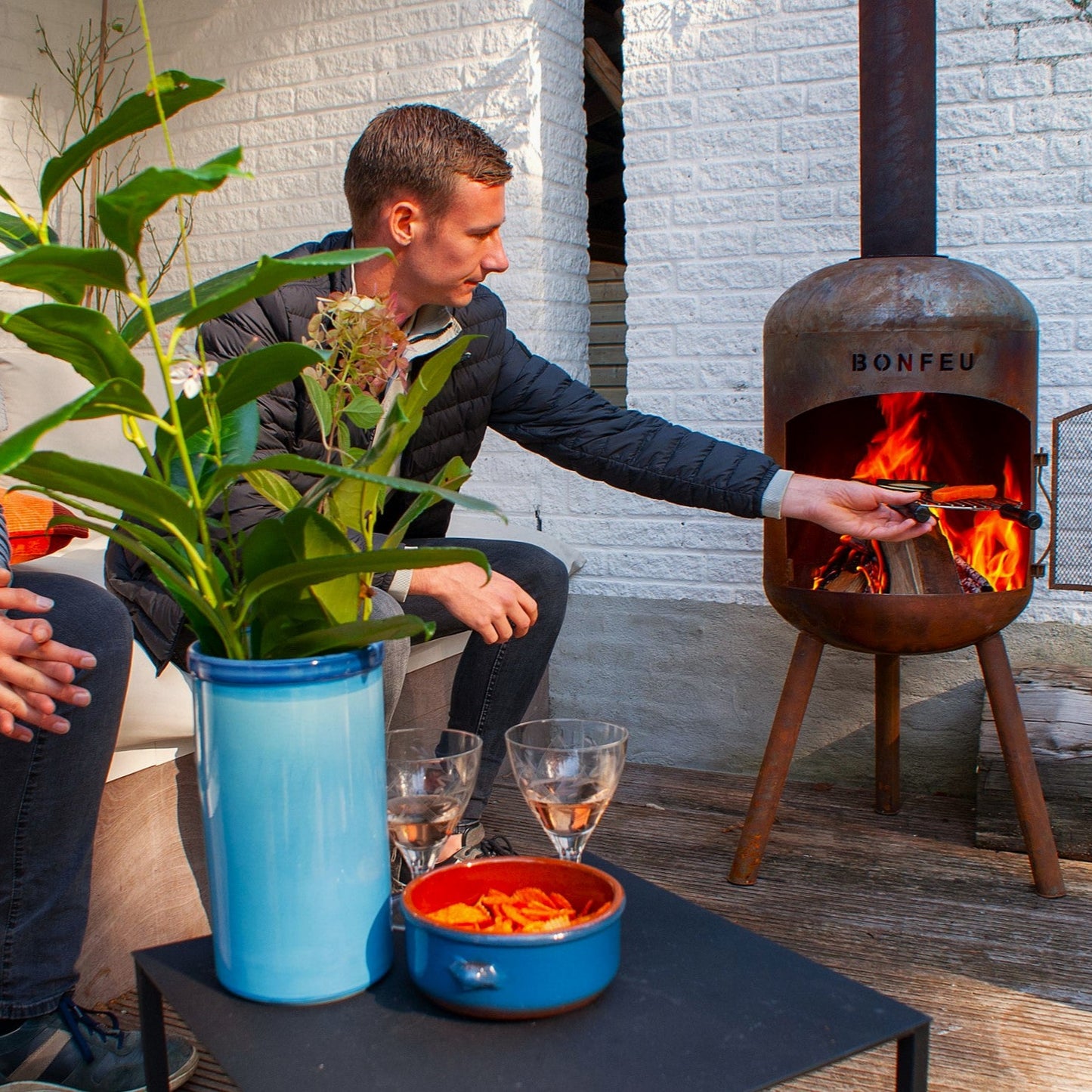 Two people sitting outdoors by a fire pit with a plant and drinks on a table.