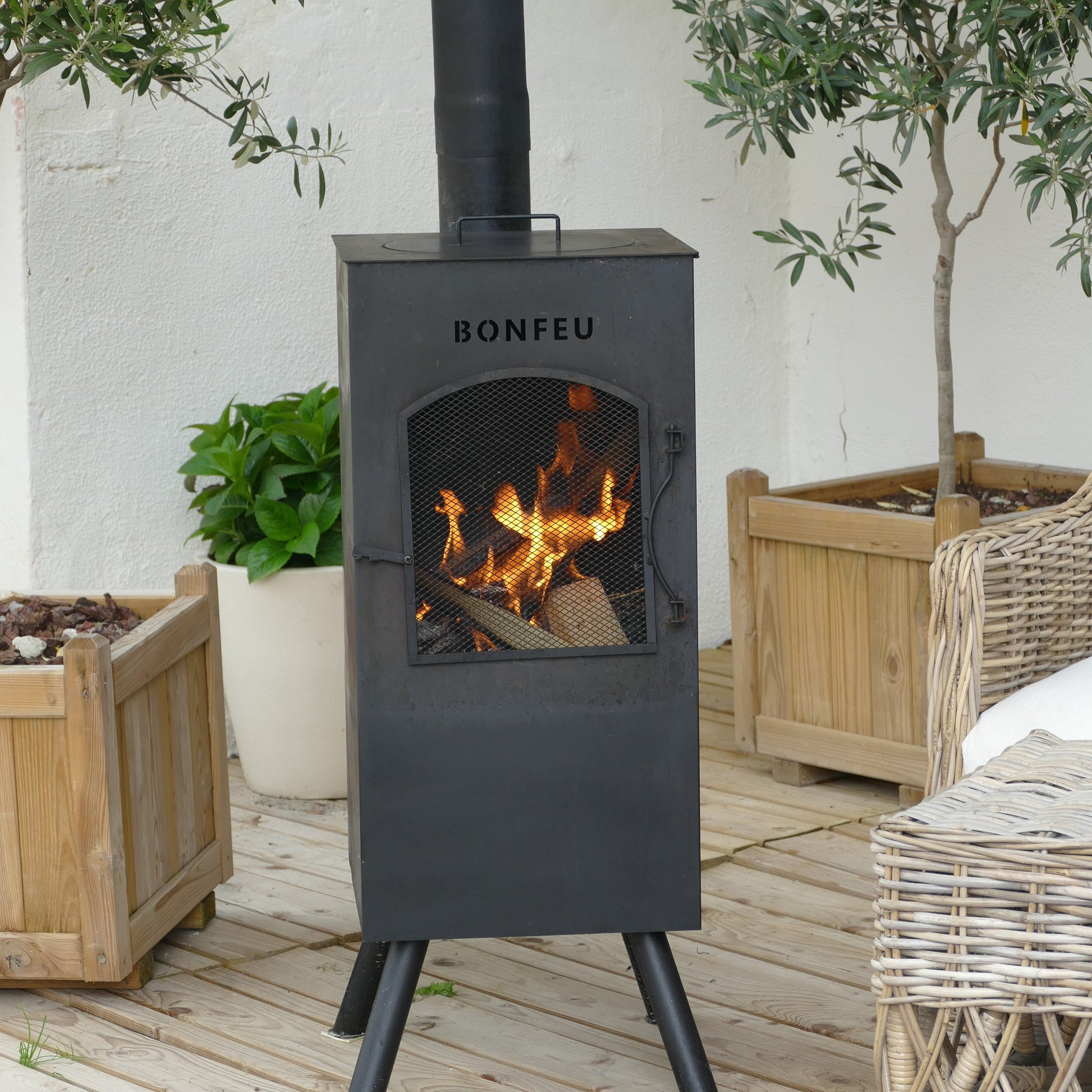 Black outdoor wood stove labeled 'BONFEU' with flames burning, surrounded by plants and furniture on a wooden deck.