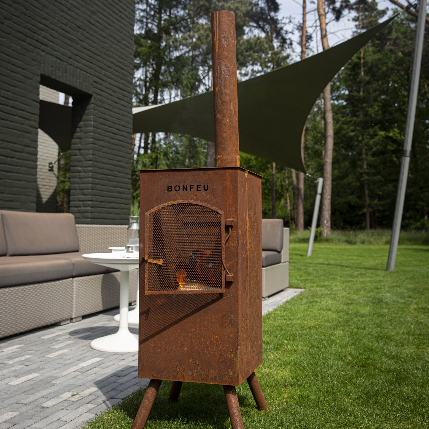 Wood-burning stove in a garden setting with a modern house and trees in the background.
