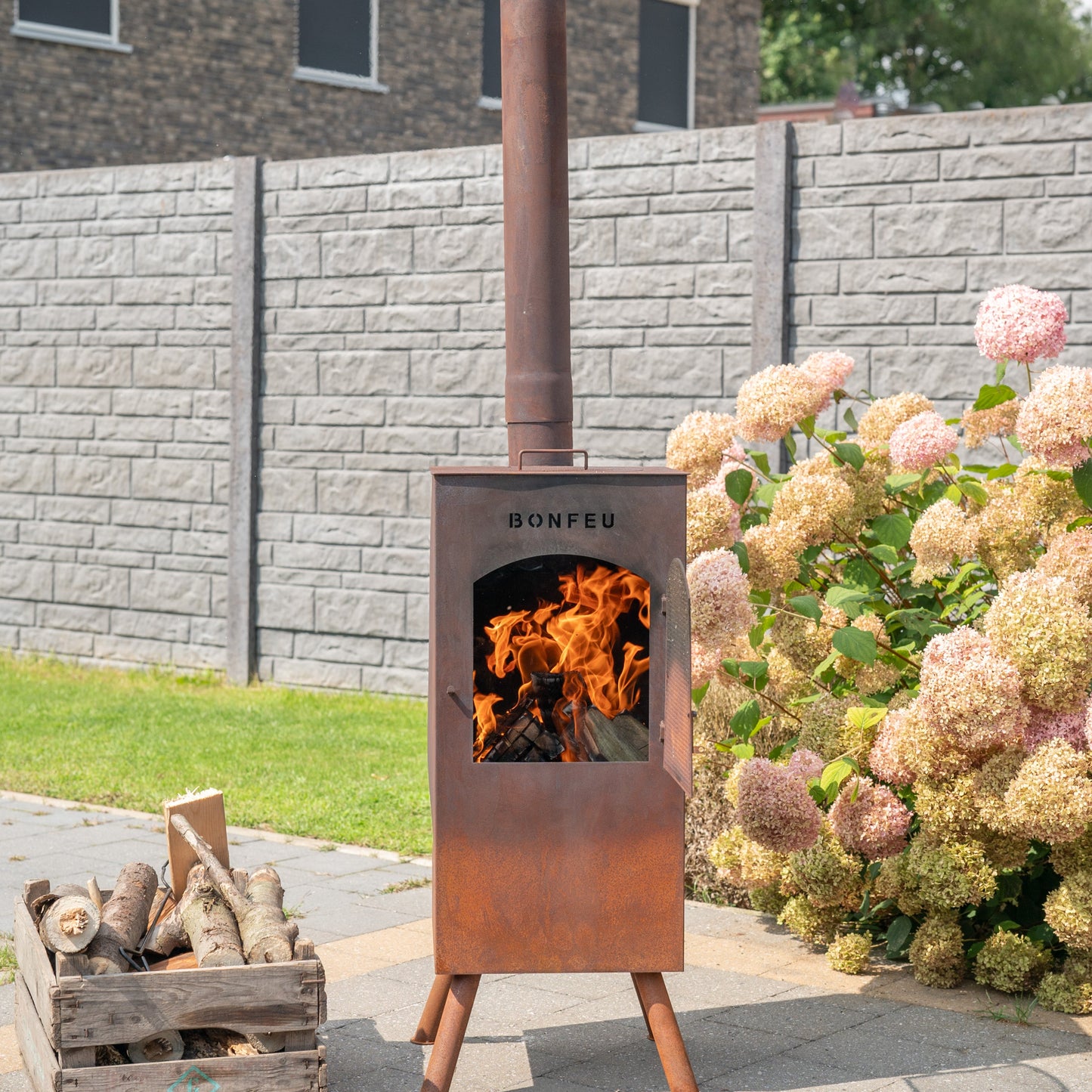 Outdoor wood-burning stove with flames, surrounded by flowers and a stone wall.