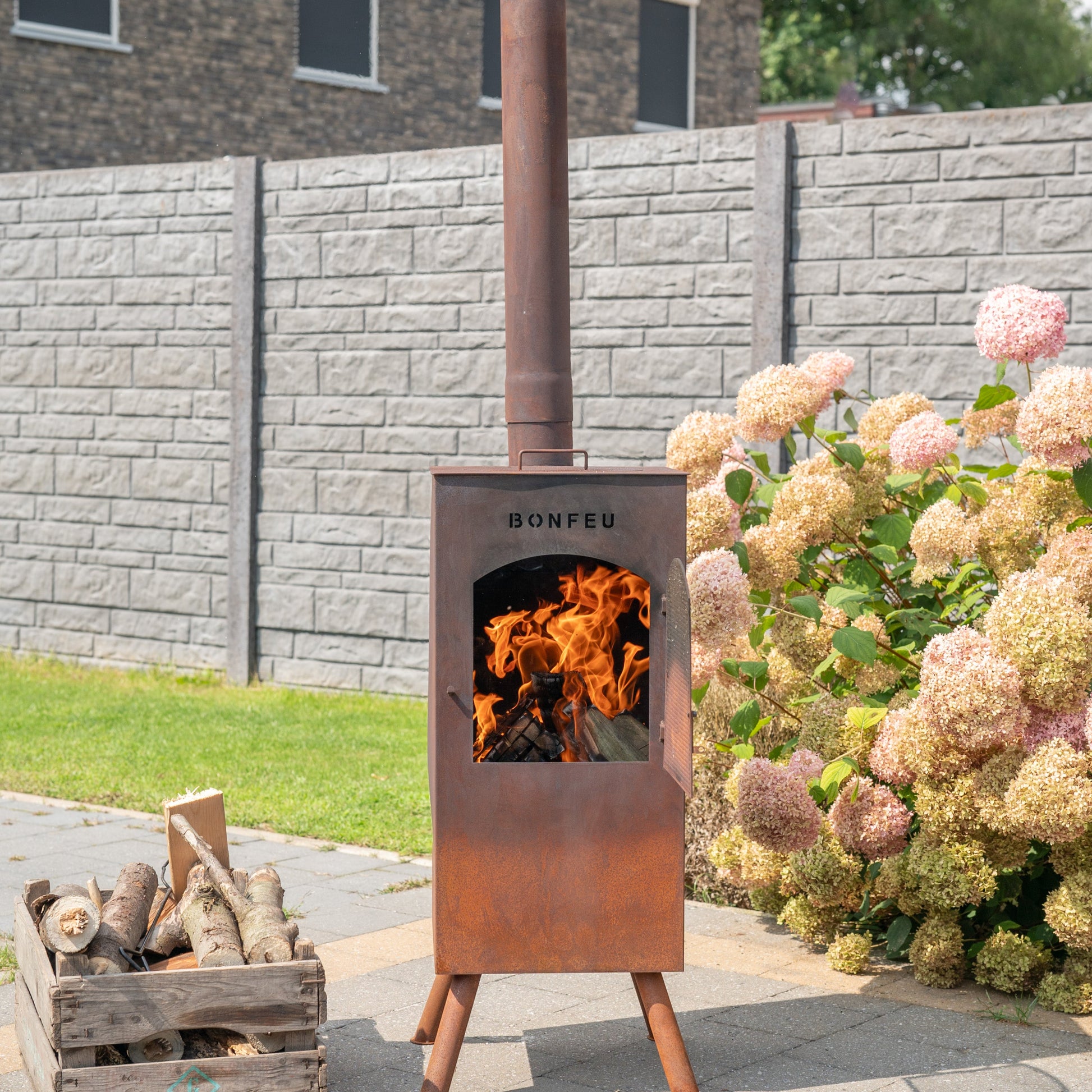 Outdoor wood-burning stove with flames, surrounded by flowers and a stone wall.