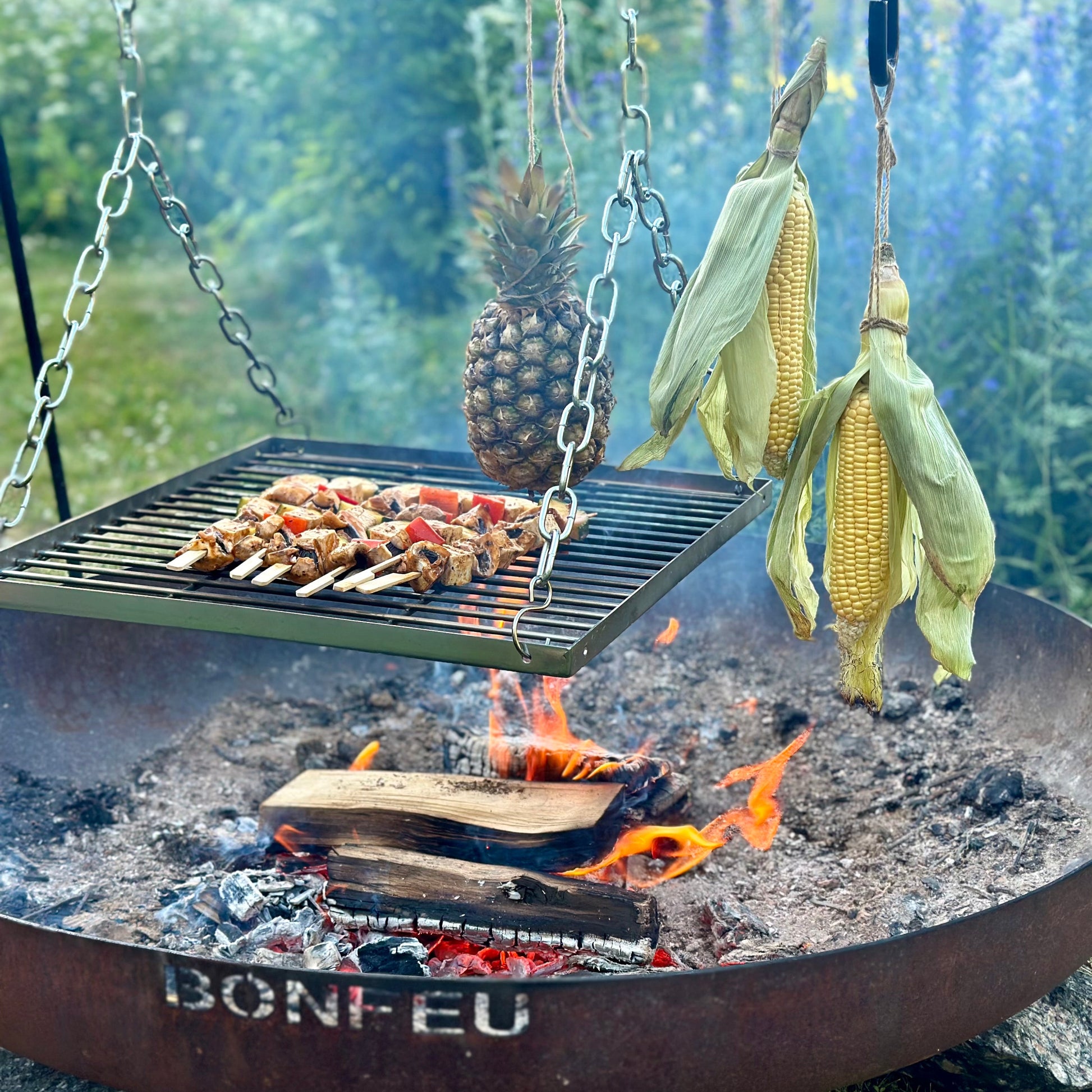 Outdoor cooking setup with skewers, pineapple, and corn on aBonfire grill.