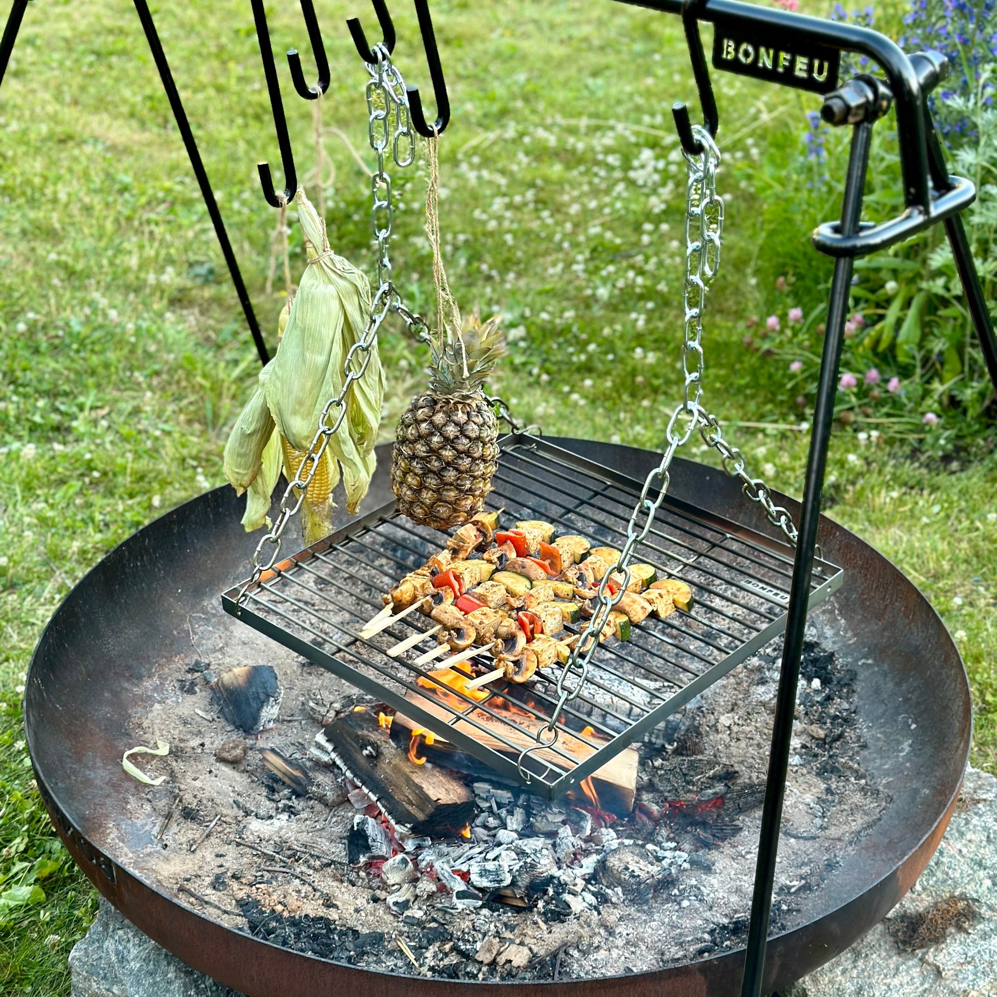 Outdoor fire pit with grill and hanging pineapple and herbs on a grassy background