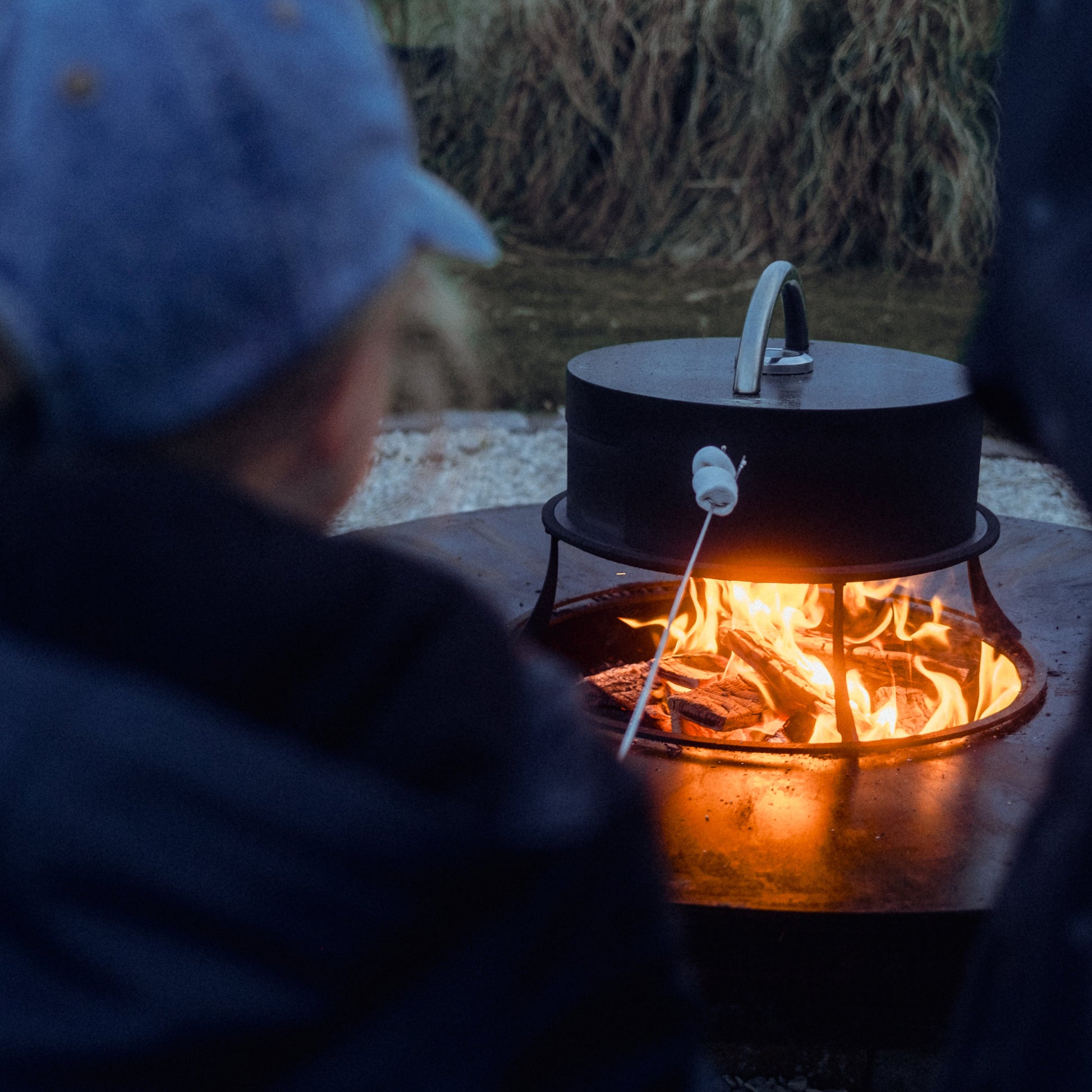 erson cooking over a fire pit with a black pot and marshmallow on a stick.