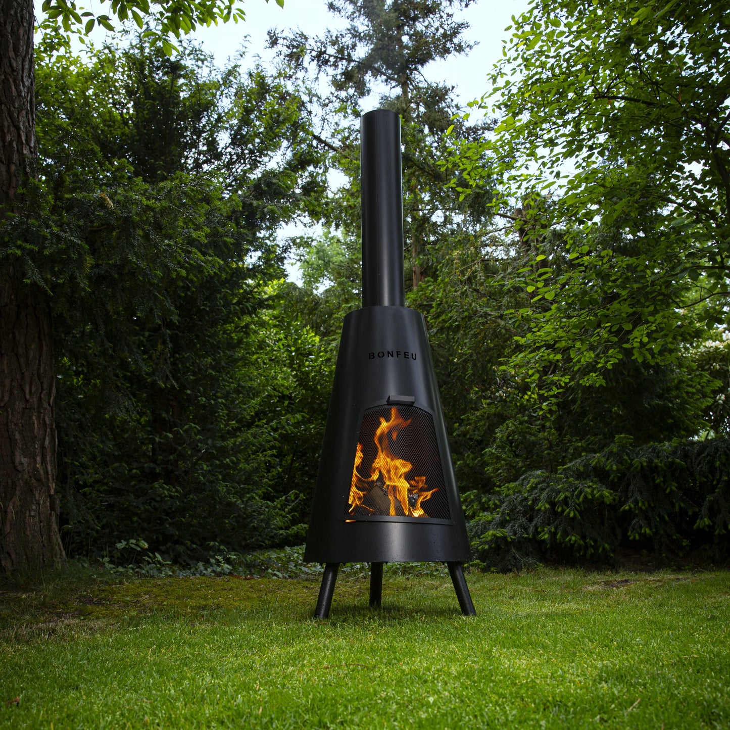 Black outdoor chiminea with flames on a grassy area with trees in the background