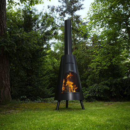 Black outdoor chiminea with flames on a grassy area with trees in the background