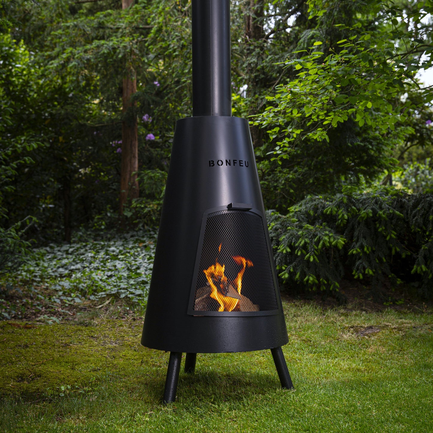 Black outdoor chiminea with flames on a grassy area with trees in the background