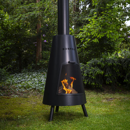 Black outdoor chiminea with flames on a grassy area with trees in the background