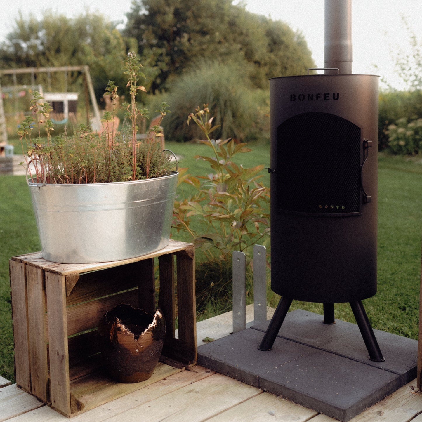 Outdoor wood stove on a wooden platform with a planter and small animal inside, set against a natural background.