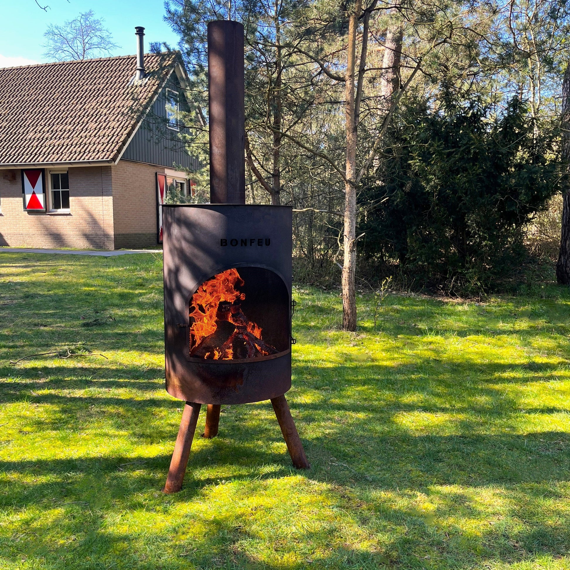 Outdoor fire pit with flames on a grassy area with trees and a building in the background