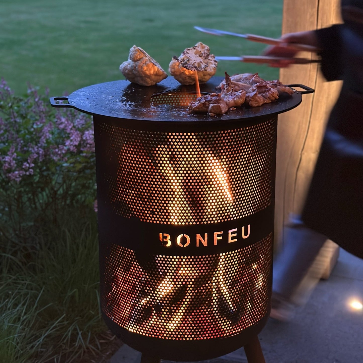 Outdoor grill with 'Bonfeu' branding being used to cook food in a garden setting.