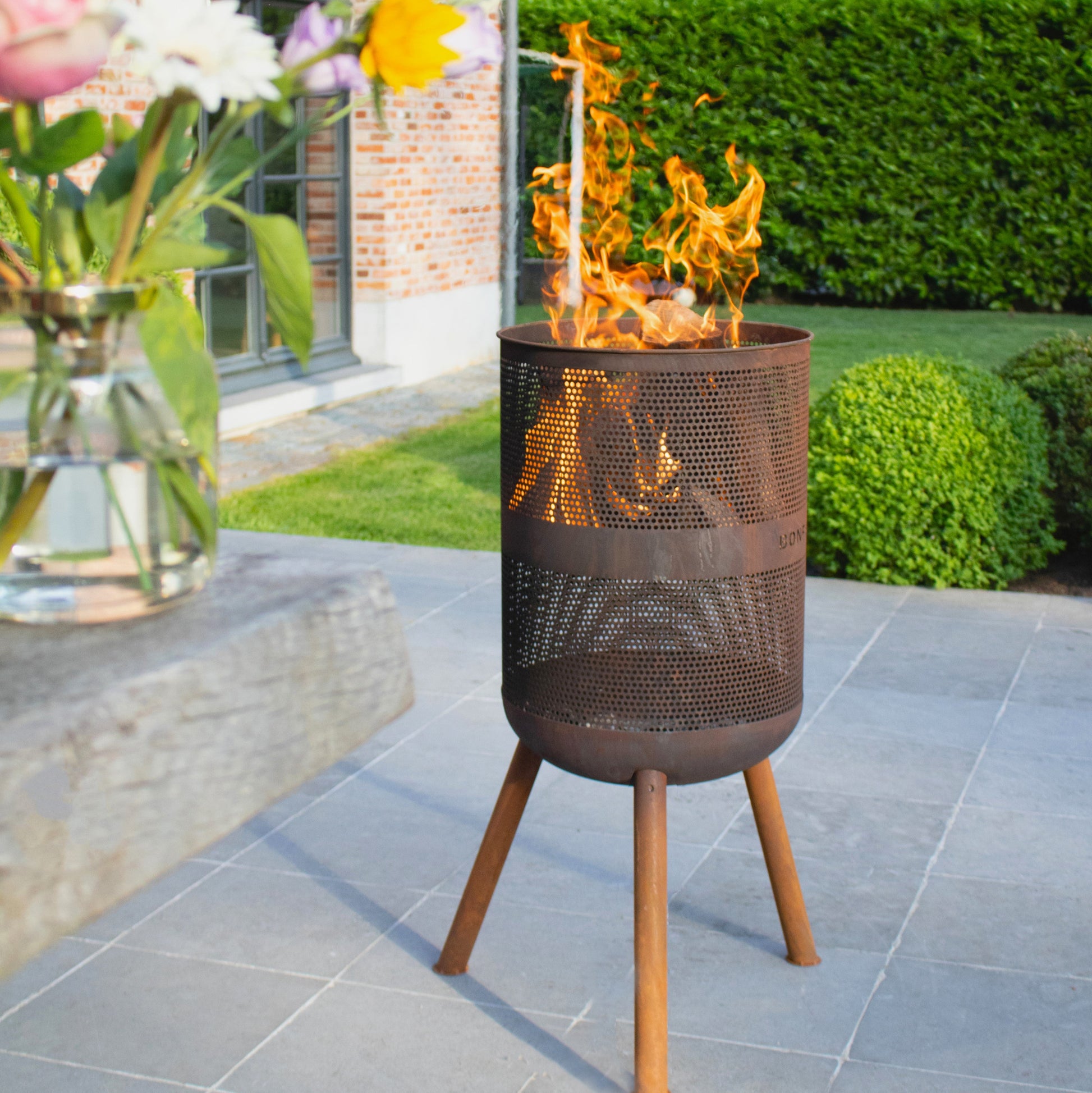 Outdoor fire pit with flames on a patio, surrounded by flowers and a building in the background.