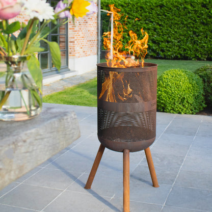 Outdoor fire pit with flames on a patio, surrounded by flowers and a building in the background.
