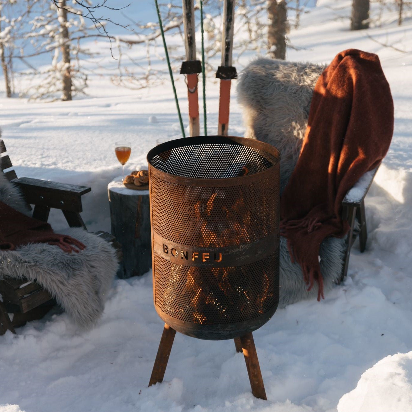 Person sitting in the snow next to a portable fire pit with skis in the background