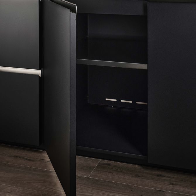 A modern black kitchen cabinet with one door open, revealing a dark interior with a middle shelf and slim silver handles, set against dark wood flooring.