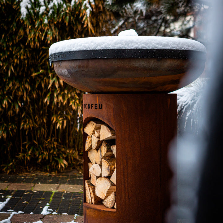 Wooden firewood holder with snow on top, surrounded by snow-covered ground and trees.
