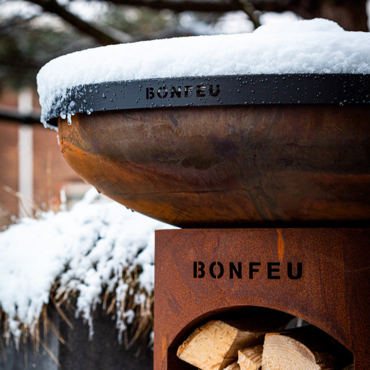 Wooden fire pit with snow on top and 'BONFEU' branding, surrounded by trees and snow.