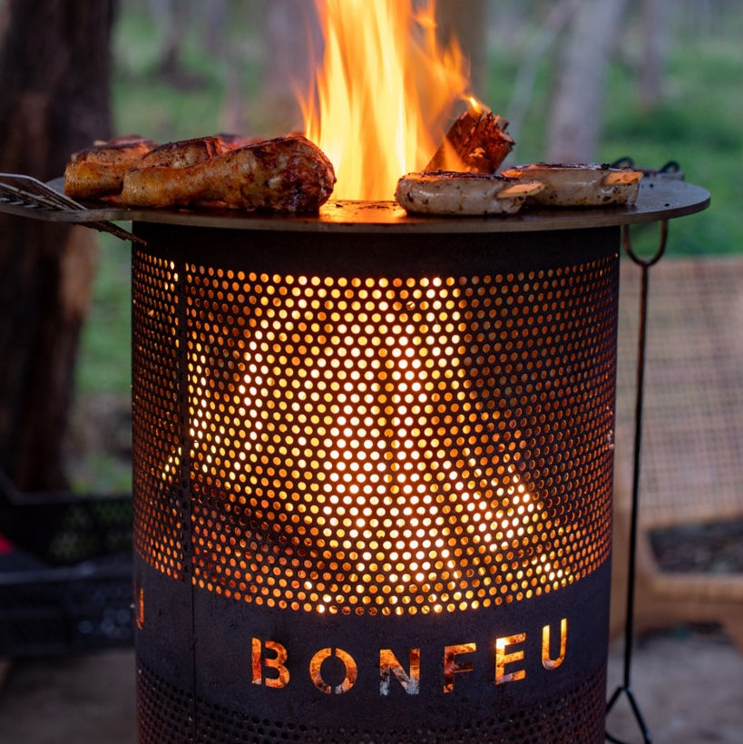 Outdoor grill with flames cooking food, branded 'BONFEU', against a natural background.