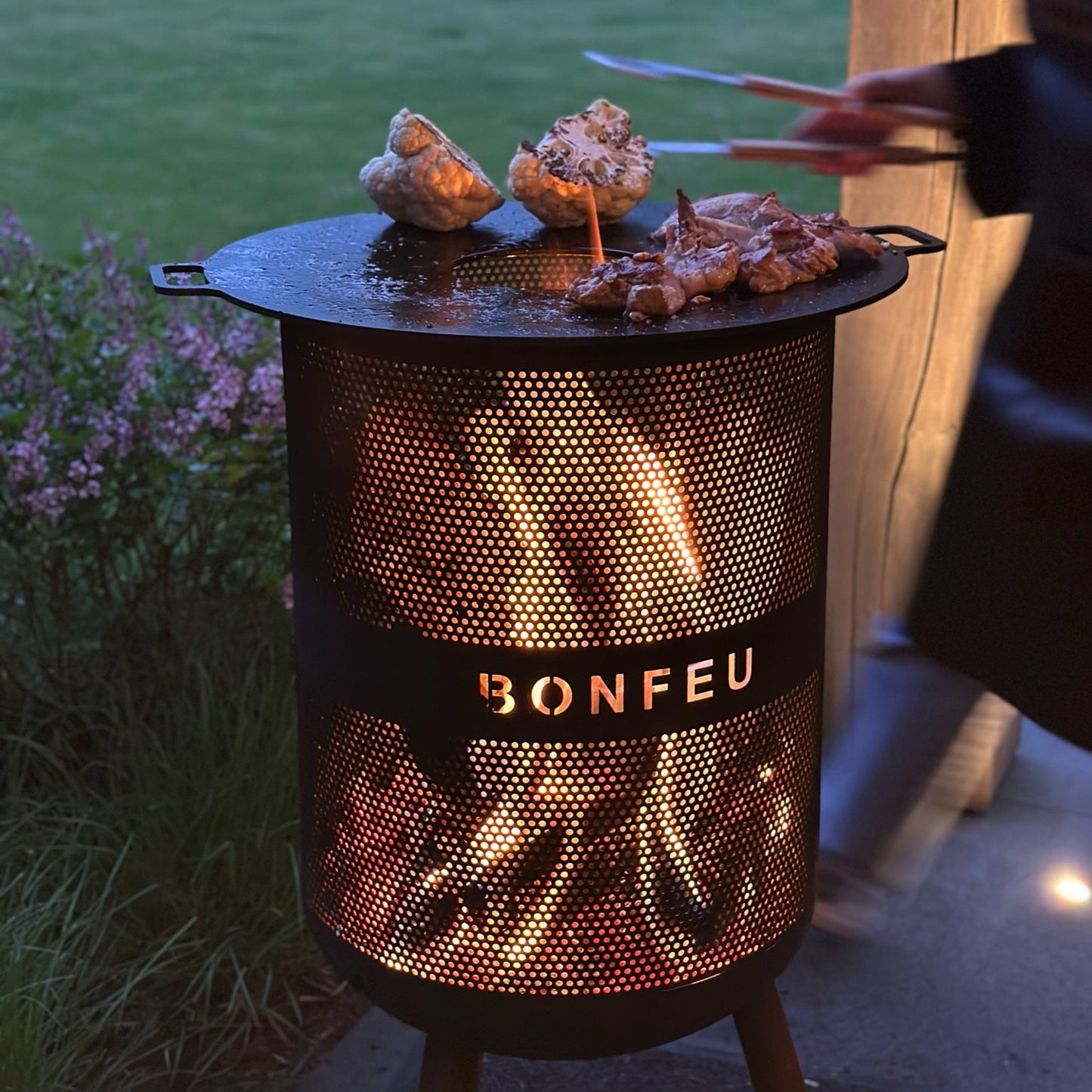 Outdoor grill with 'Bonfeu' branding being used to cook food in a garden setting.