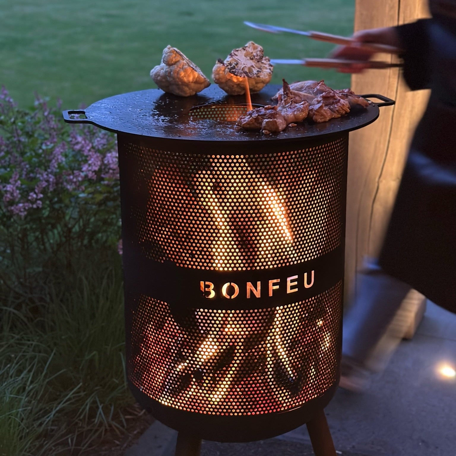 Outdoor grill with 'Bonfeu' branding being used to cook food in a garden setting.