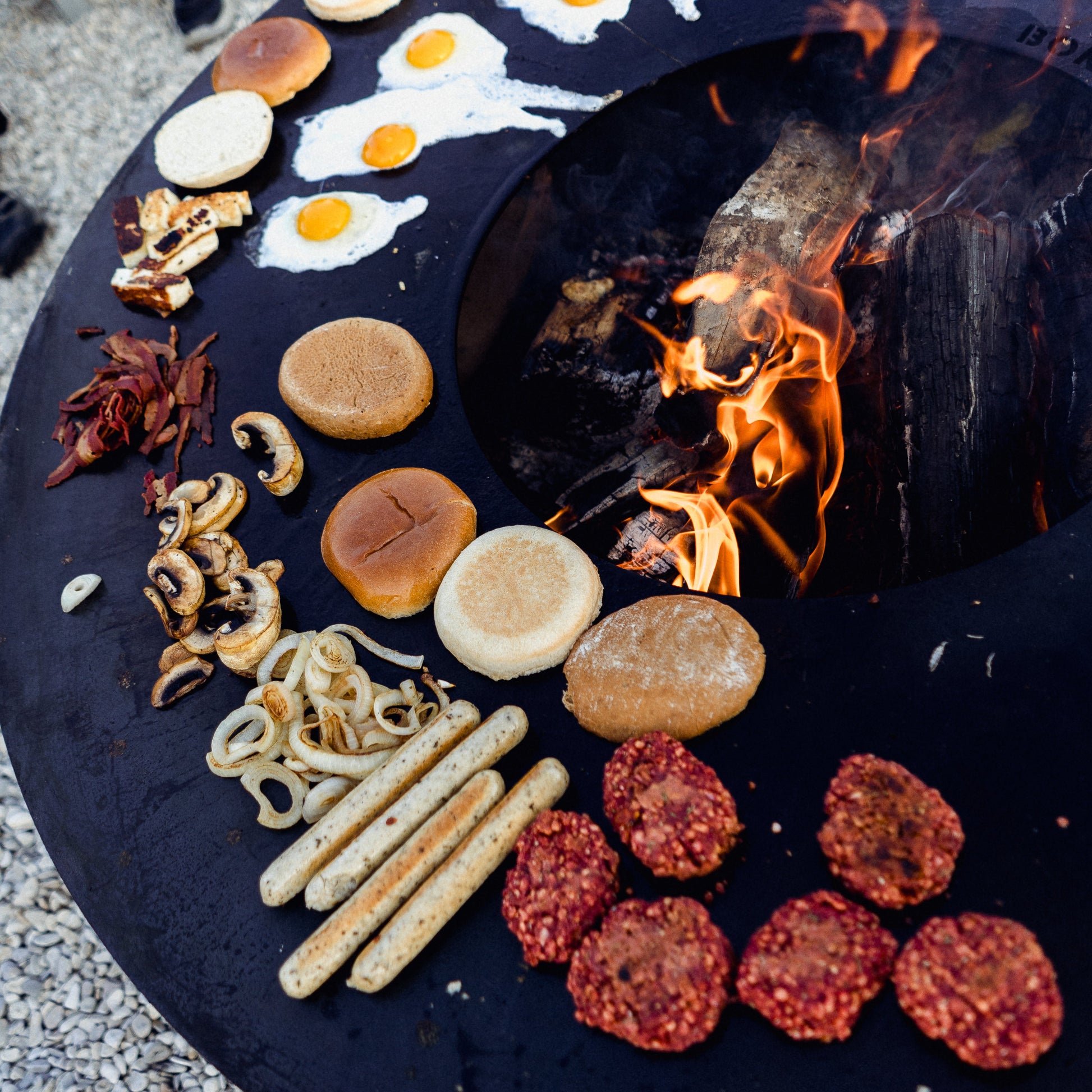Fire pit with food items like buns, sausages, and bread around a fire.