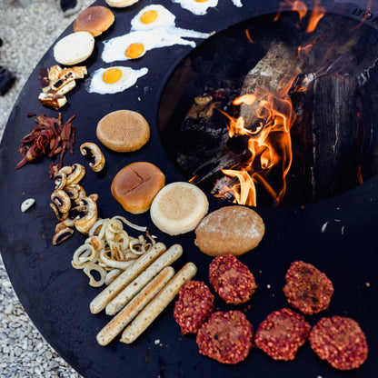 Fire pit with food items like buns, sausages, and bread around a fire.