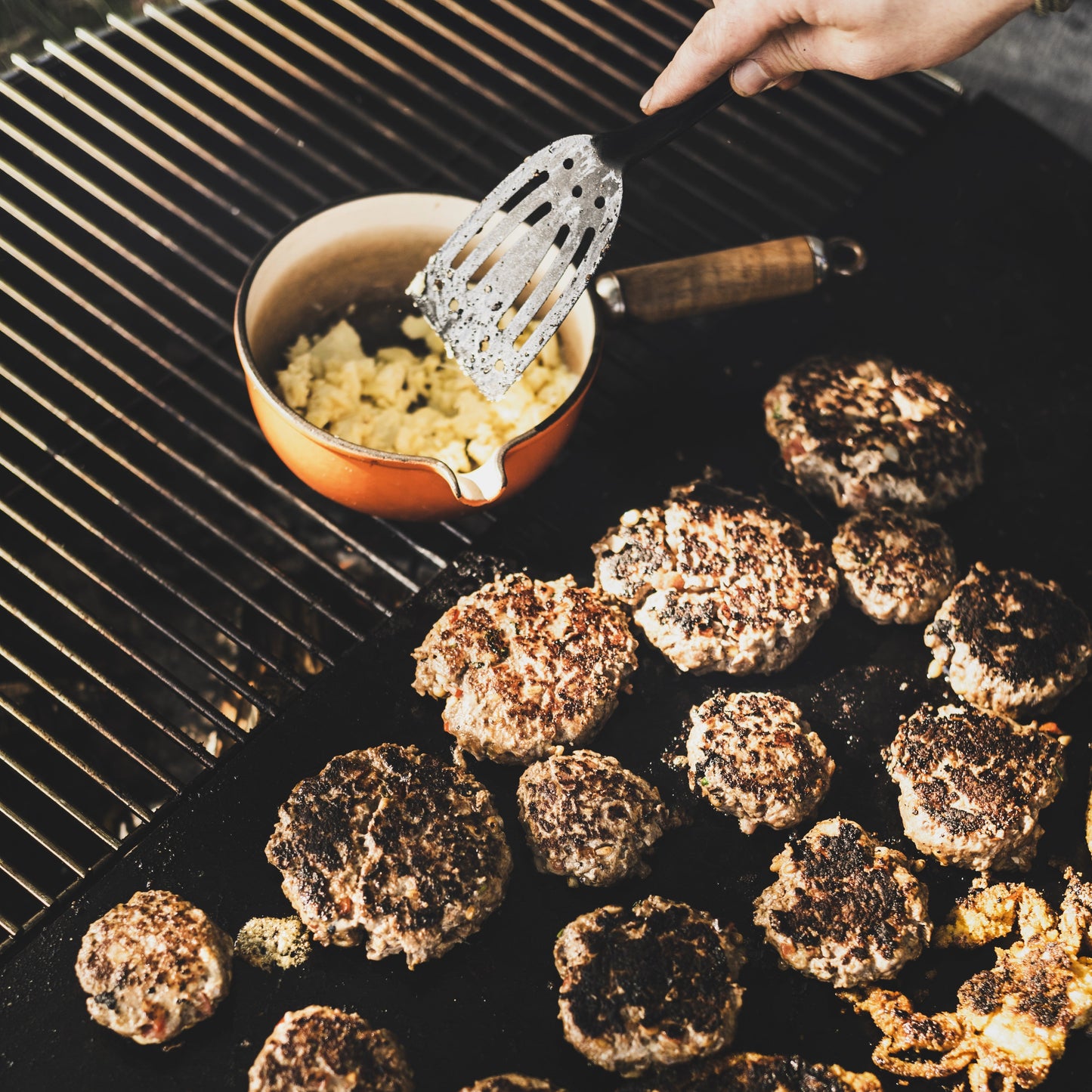 Grilled burgers on a barbecue with a hand holding a spatula and bowl of condiments.