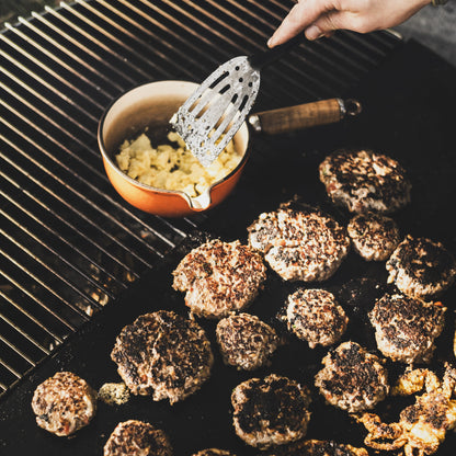 Grilled burgers on a barbecue with a hand holding a spatula and bowl of condiments.
