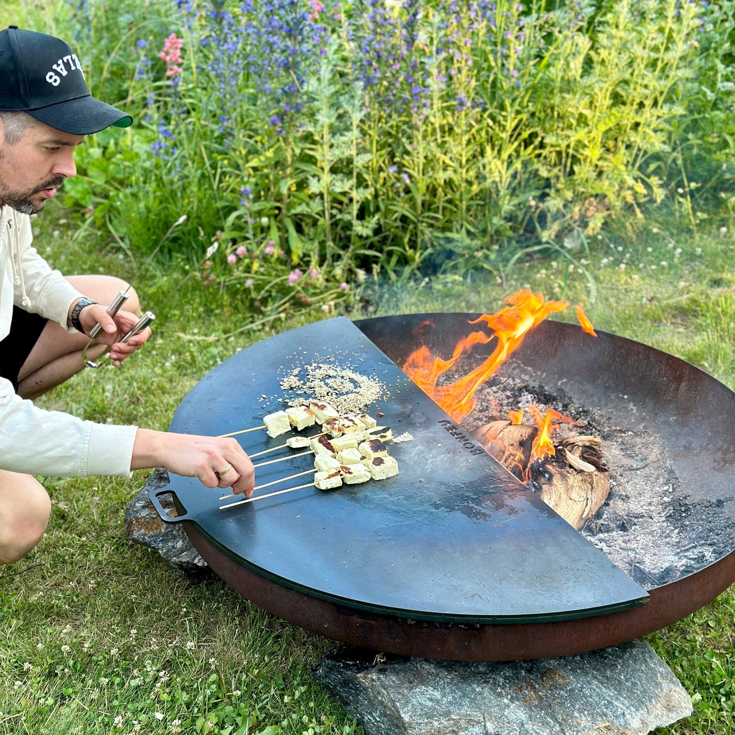 Person grilling over a fire pit in a natural setting with flowers and grass.